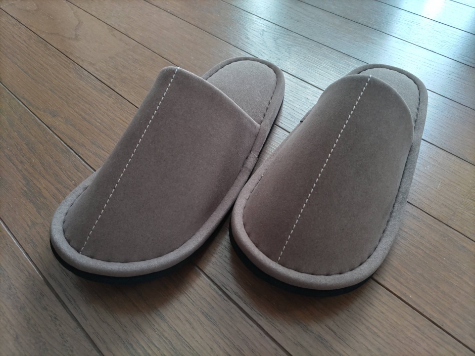 木目の床の上に並べられたブラウンの『匠のスリッパ』を正面やや斜めから撮影した写真。丸みのあるつま先や縫い目の丁寧さがはっきり見える構図。