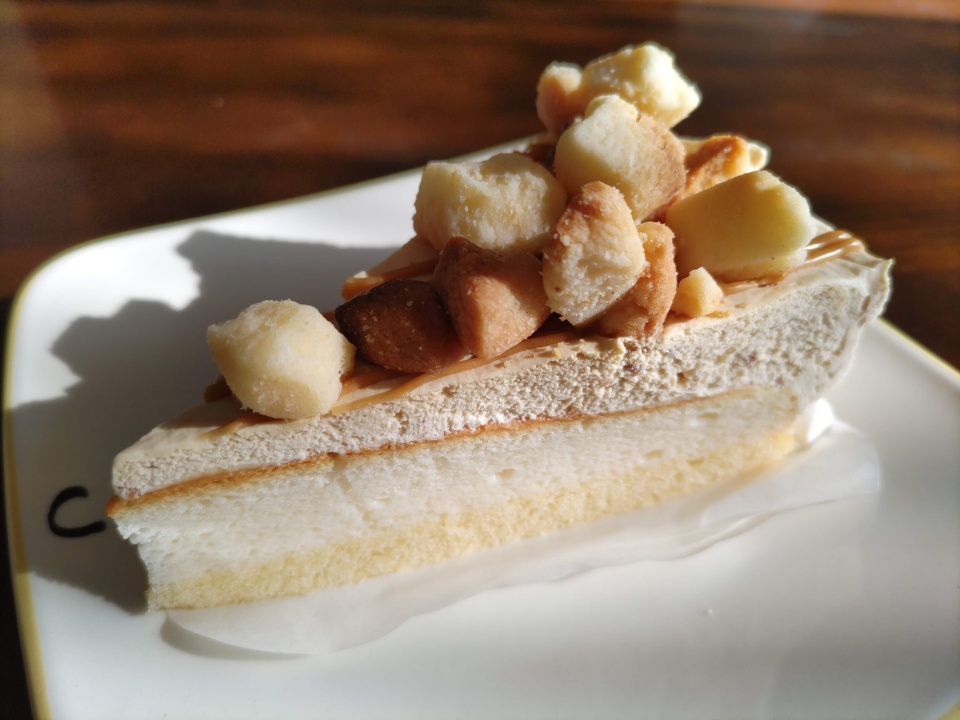 太陽の光を受けて輝くヤマザキのキャラメルチーズケーキ。ふわふわの白いスポンジの上に、淡いキャラメル色のチーズクリームが重なり、さらにその上にクランブルがごろっと乗っている。自然光によってケーキの影が皿の上に落ち、温かみのある午後の雰囲気が感じられる近接ショット。