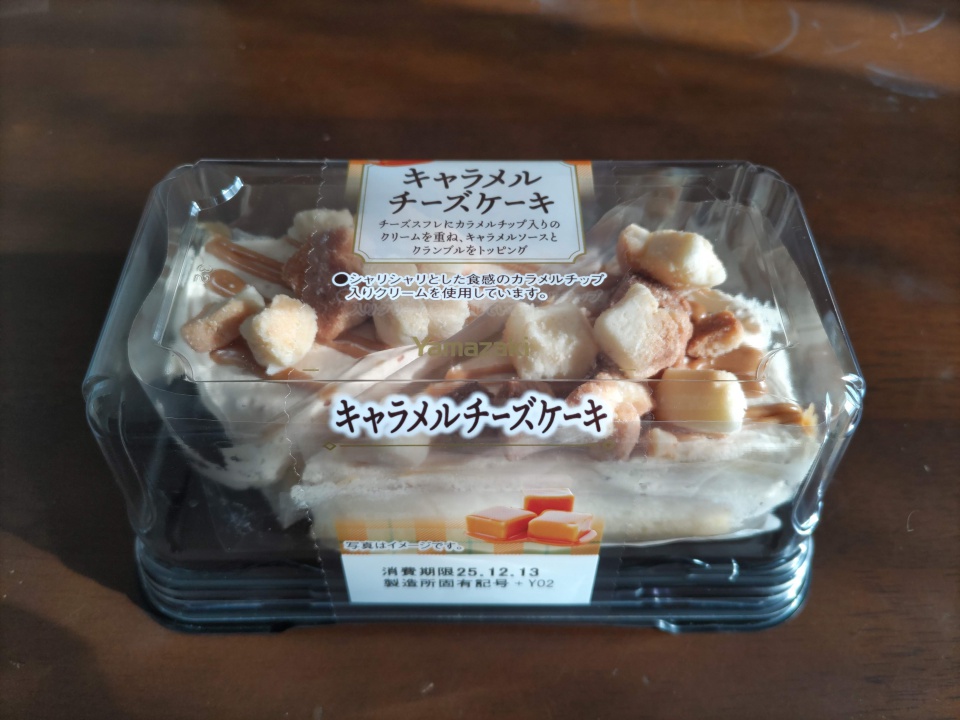 透明なプラスチックケースに入ったヤマザキのキャラメルチーズケーキ。表面にはクランブルやキャラメルソースがトッピングされており、パッケージのラベルには商品名と説明文が印字されている。自然光が横から差し込み、ケースの表面に淡い反射と影ができている様子が写っている。