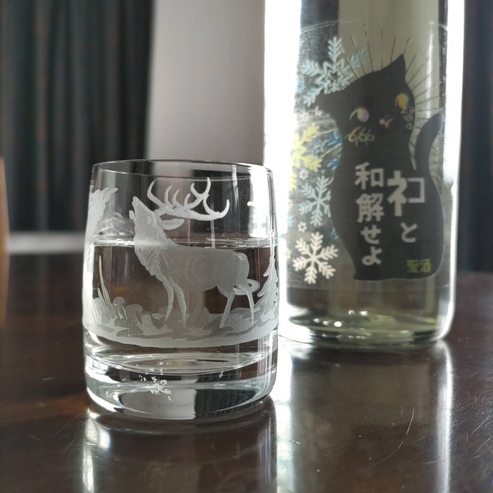 日本酒『ネコと和解せよ』の720ml瓶と、鹿の模様が彫られたガラスのお猪口に注がれた日本酒を並べて撮影した写真。瓶には黒猫のイラストと雪の結晶が描かれた冬仕様のラベルが貼られており、柔らかな室内光の中でテーブルに静かに佇んでいる。