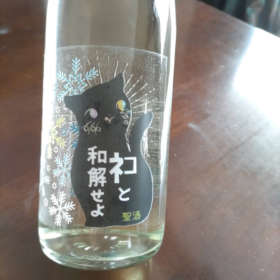 日本酒『ネコと和解せよ』の瓶をテーブルの上で撮影した写真。黒い猫のシルエットと雪の結晶が描かれた冬仕様のラベルが大きく写っており、文字がはっきり見えるように自然光が差し込む構図になっている。