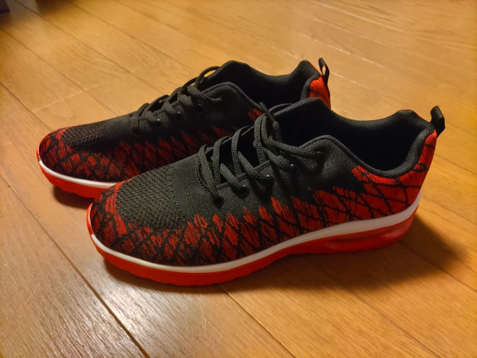 木目調のフローリングの上に並べて置かれた、ブラックとレッドの配色が特徴的なスニーカー。アッパーはナイロン素材で、赤い模様がアクセントになっており、厚みのある赤いソールがスポーティーな印象を与えている。