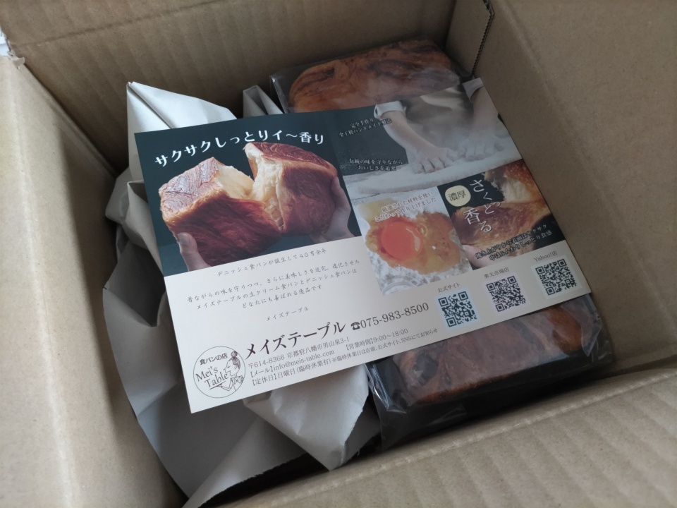 高級スイーツデニッシュ栗1斤が入った段ボール箱を開封した状態の写真。緩衝材に包まれたデニッシュと、商品説明が掲載されたメイズテーブルのリーフレットが同梱されている様子。