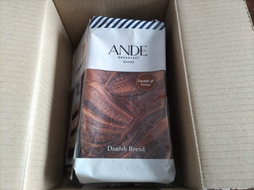 アンデ「ショコラーデデニッシュ」1斤が入った段ボール箱を開けた直後の写真で、箱の中央に白いパッケージのデニッシュ食パンが収まっており、「ANDE BREAKFAST KYOTO」「Danish Bread」「ショコラーデ」の文字とマーブル模様のパンの写真が見える様子を写している。