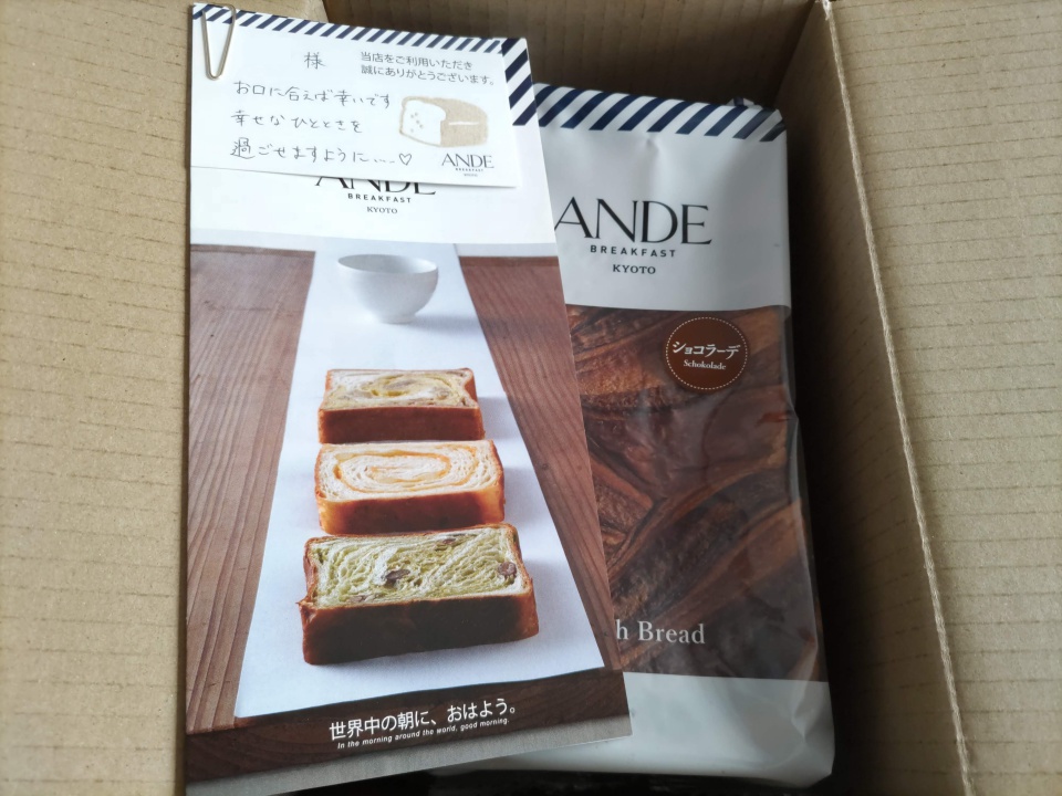 アンデのデニッシュ食パンが入った段ボール箱の中を写した写真で、右側に「ANDE BREAKFAST KYOTO」と表示されたショコラーデデニッシュのパッケージ、左側にスライスされたデニッシュの写真が掲載された商品案内の冊子が一緒に収まっている様子が分かる。