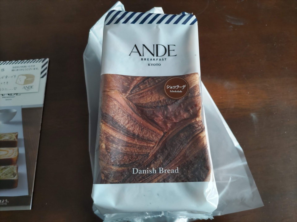 アンデ「ショコラーデデニッシュ」1斤を段ボール箱から取り出した直後の写真で、木目のテーブルの上に白いパッケージのデニッシュ食パンが置かれ、「ANDE BREAKFAST KYOTO」「ショコラーデ」「Danish Bread」の文字とチョコレートデニッシュのマーブル模様がはっきり見える様子を写している。