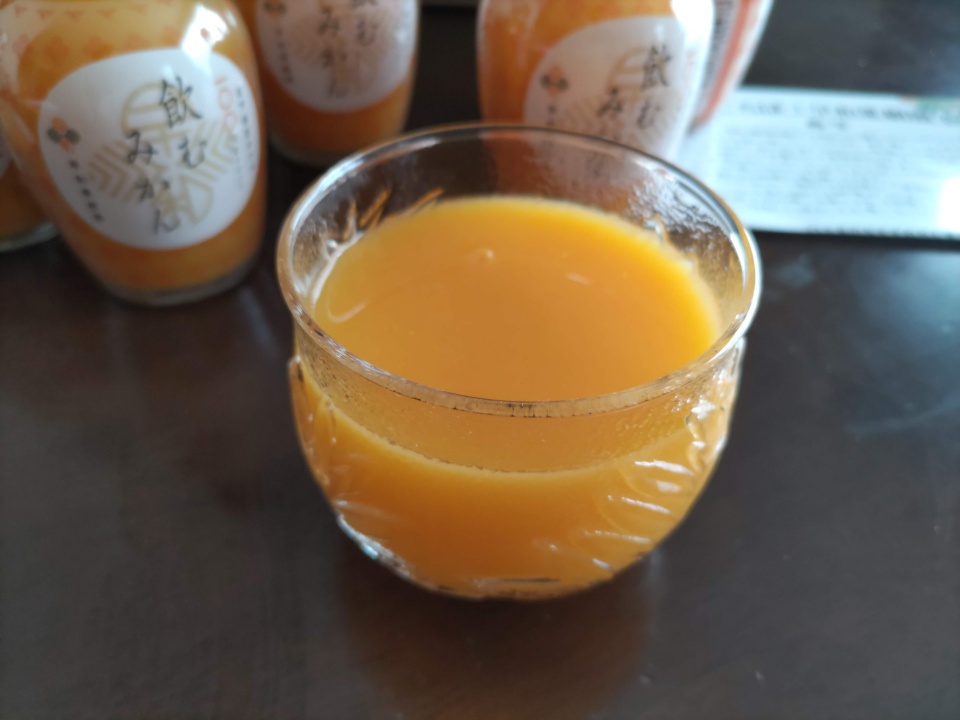 飲むみかんをグラスに注いだ様子。濃いオレンジ色のとろみのあるジュースがガラスのコップに入っており、背景には早和果樹園の「飲むみかん」のボトルが並んでいる。