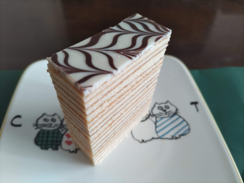 志布志金山バームの一切れを白い皿に盛り付けた写真。16層にも重なったしっとり生地の断面と、上部の白とチョコの矢羽根模様のコーティングが美しく際立っている。皿には猫のイラストが描かれている。