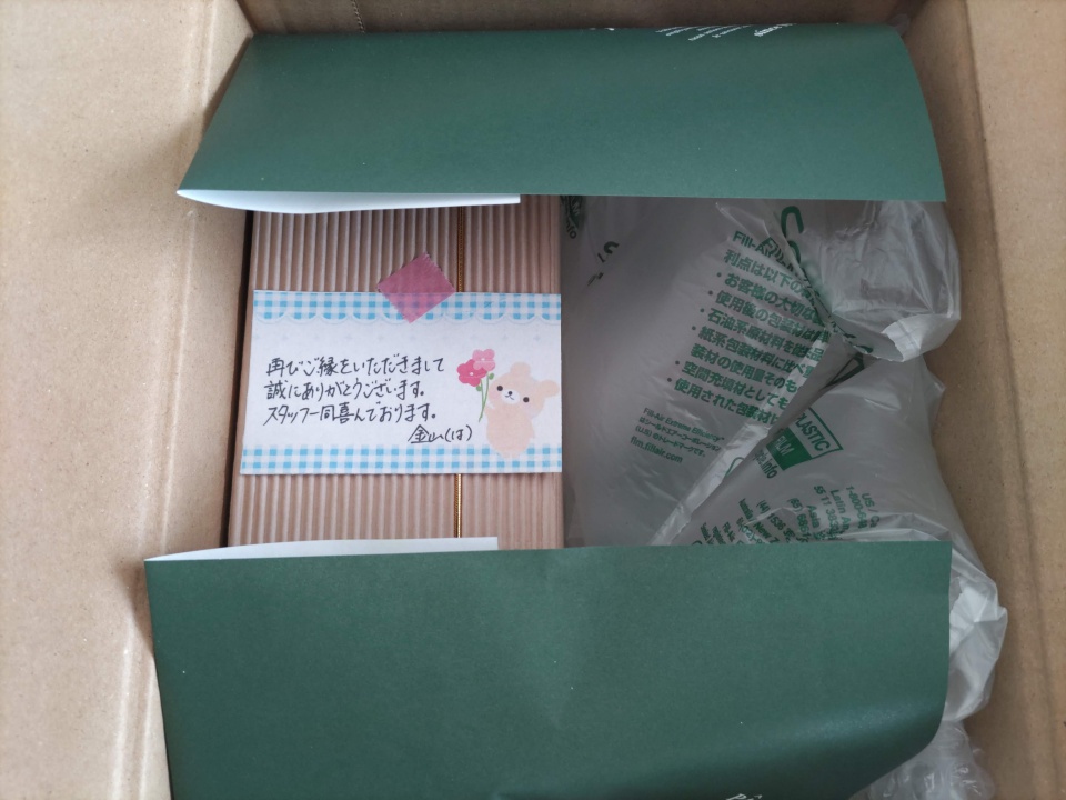 ゆうパックで届いた志布志金山バームの外箱を開封した直後の写真。緑色の包装紙に包まれた箱と緩衝材が見え、中央には「再びご縁をいただきまして誠にありがとうございます。スタッフ一同喜んでおります。」と書かれた手書きメッセージカードが添えられている。