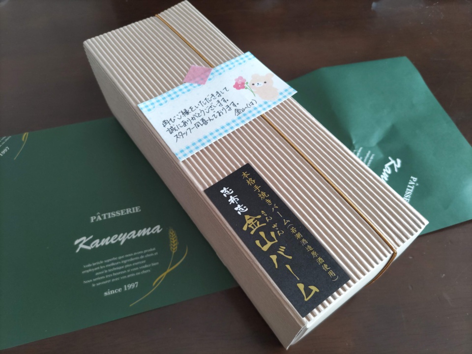 志布志金山バームの化粧箱をテーブルの上に置いて撮影した写真。縦筋の入った上品な箱に「金山バーム」と書かれた黒い帯がかかり、上には手書き風のメッセージカードが添えられている。背景には緑色の包装紙が広げられている。