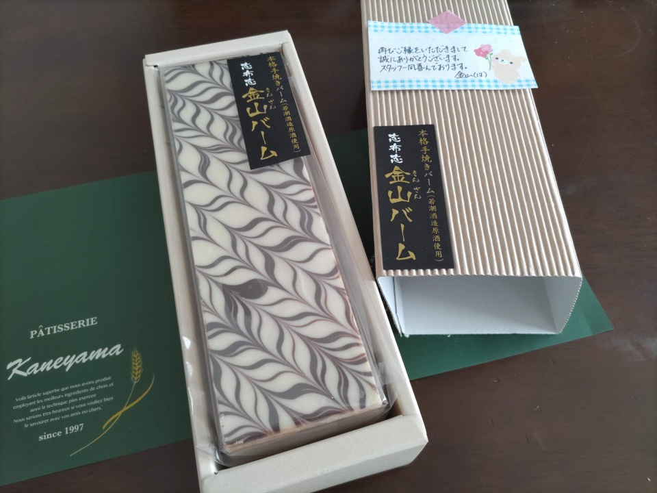 志布志金山バームの化粧箱のふたを開けた状態の写真。箱の中には、白とチョコの矢羽根模様が美しいバームクーヘンが透明フィルムに包まれて収められている。横にはふたとメッセージカードが置かれ、背景には緑色の包装紙が広がっている。