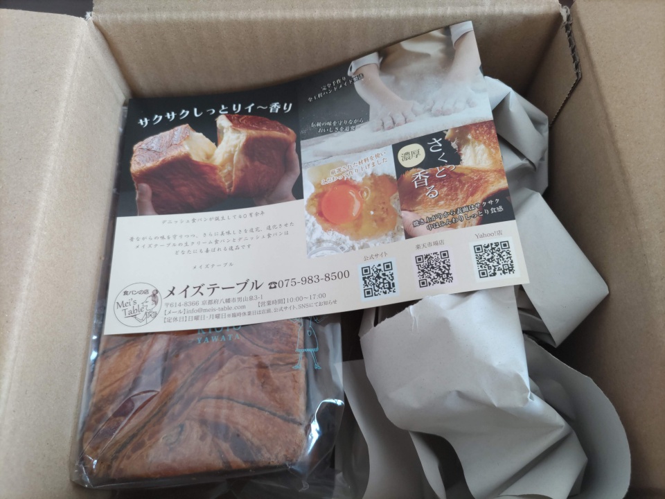 段ボール箱を開封した状態で、緩衝用の紙に包まれた桜餡デニッシュと、メイズテーブルの紹介チラシが同梱されている様子。