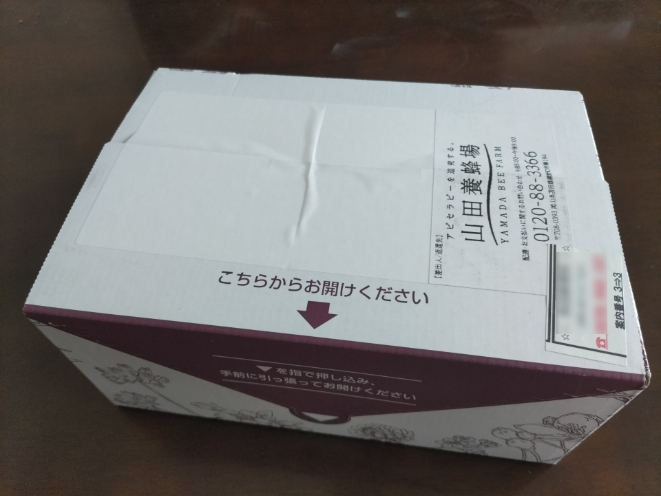 山田養蜂場のはちみつバウムが宅配で届いた状態のダンボール箱の写真。未開封の箱の上面には送り状と「こちらからお開けください」という案内が表示されている。