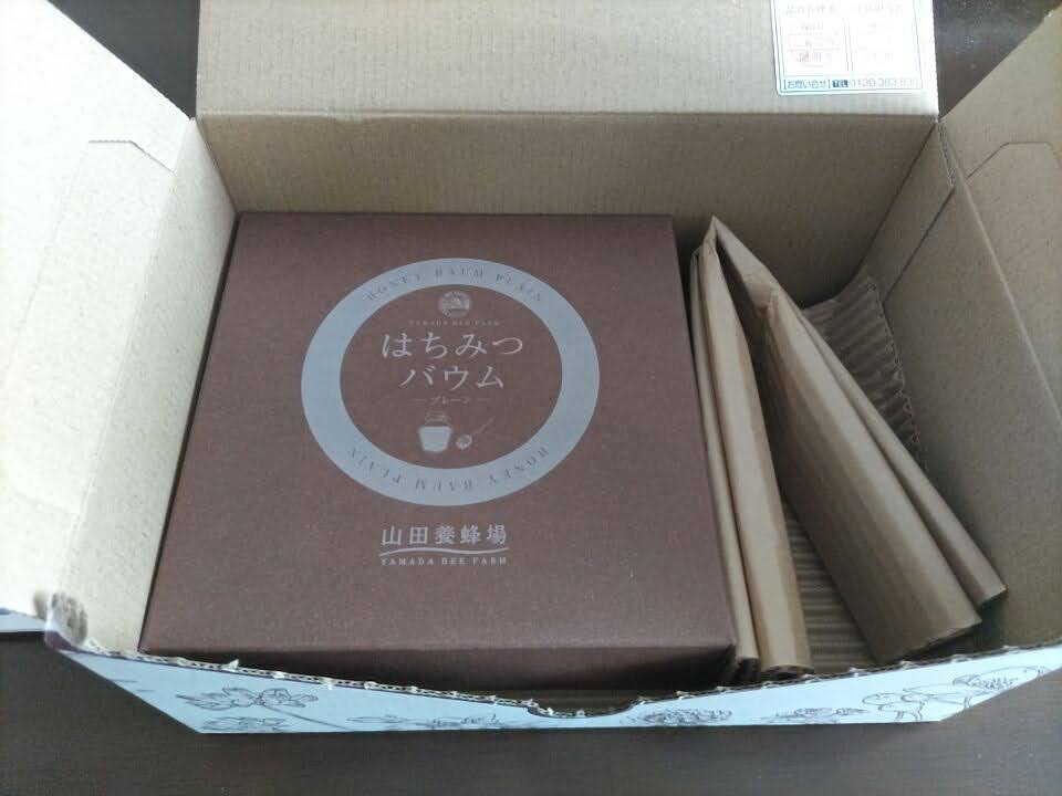 開封したダンボール箱の中に、山田養蜂場の「はちみつバウム」の化粧箱が収められている様子を写した写真。筒状の緩衝材や納品書を取り除いた状態で、商品パッケージがはっきり確認できる。