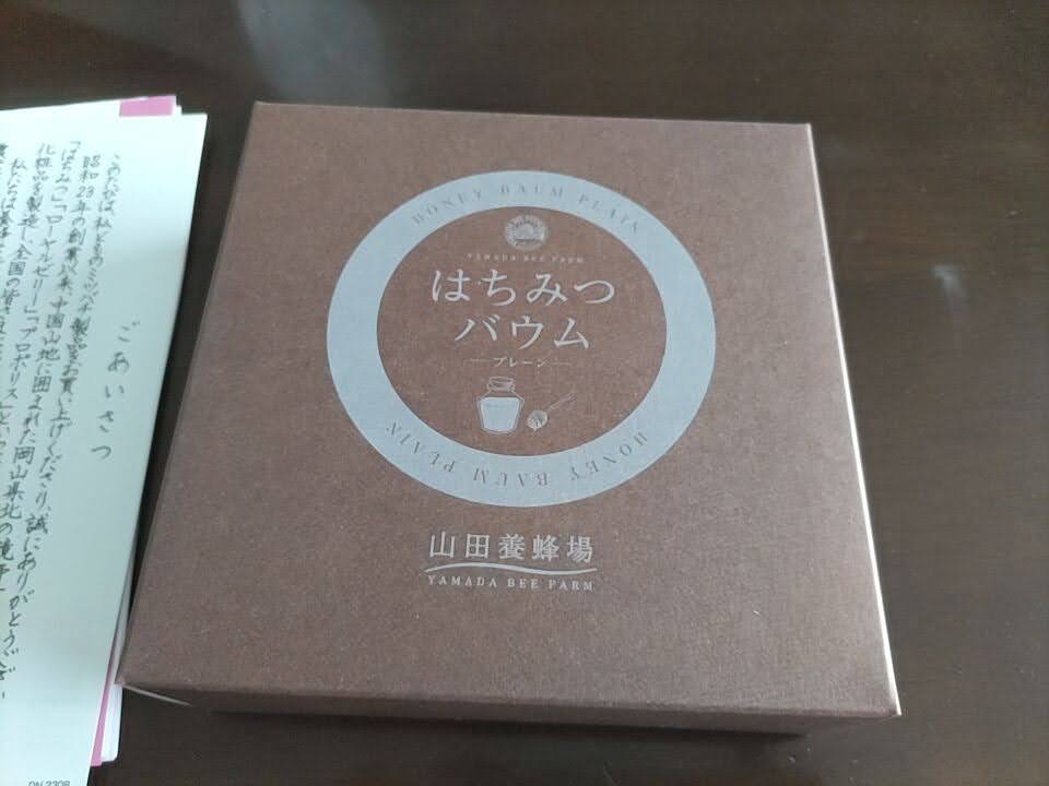 山田養蜂場の「はちみつバウム」の化粧箱を段ボール箱から取り出し、テーブルの上に置いた写真。落ち着いたブラウンのパッケージに商品名とブランドロゴがデザインされている。