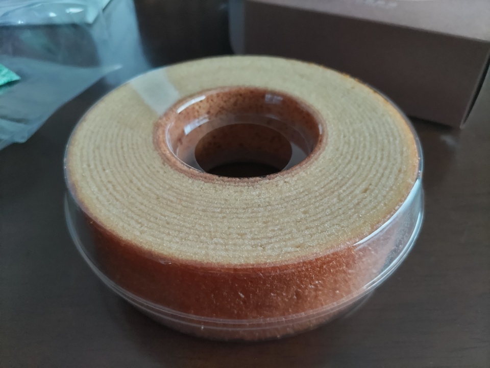 袋から取り出した山田養蜂場のはちみつバウムを、透明なプラスチックカバーに入れた状態でテーブルに置いた写真。しっとりとした層が重なった円形のバウムクーヘンが確認できる。