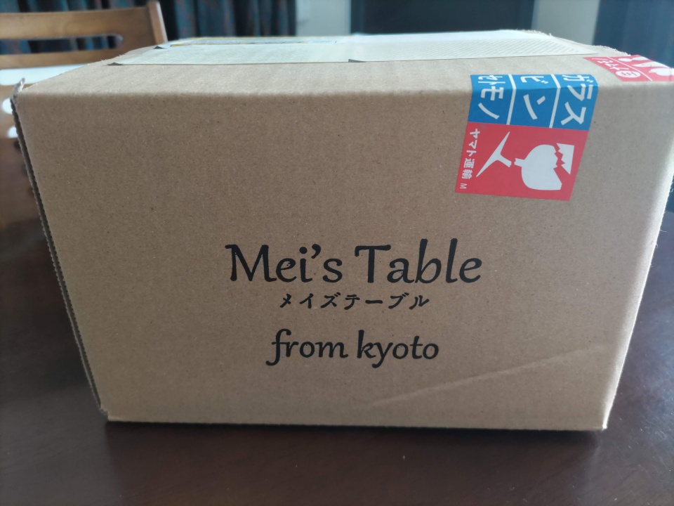 「Mei’s Table（メイズテーブル）from kyoto」と印字された茶色のダンボール箱。側面には「ワレモノ」と書かれた取扱注意シールが貼られ、宅配便で届いた様子がわかる。